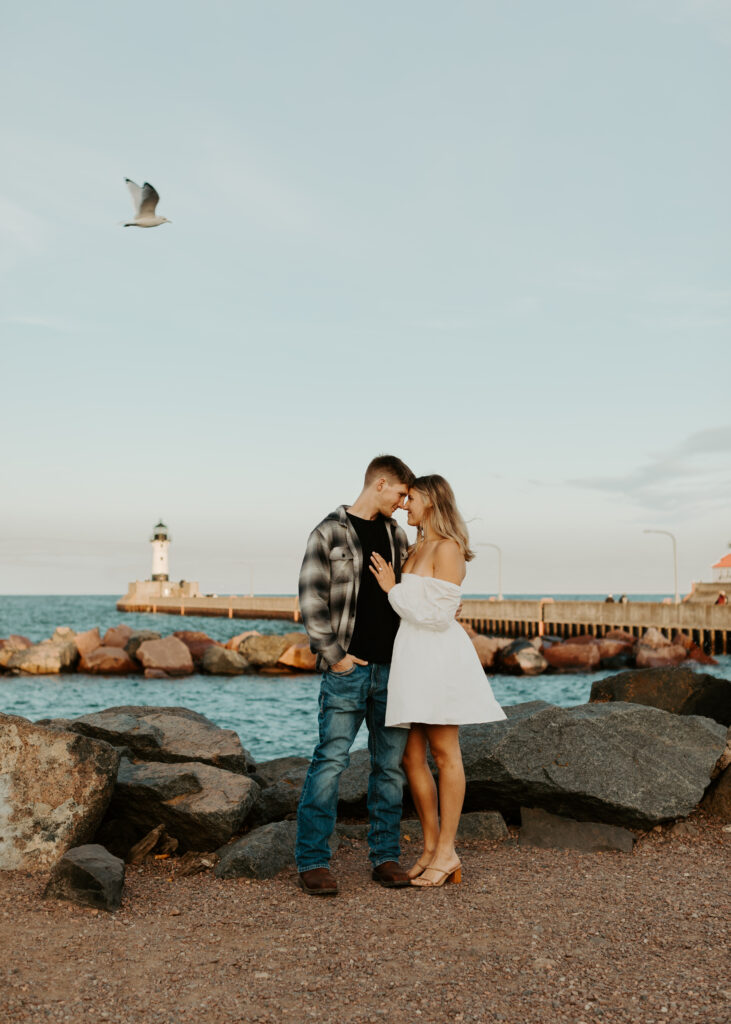 Couple stands in front of Lake Superior as a seagull flies above in Canal Park Duluth Minnesota during engagement photo session